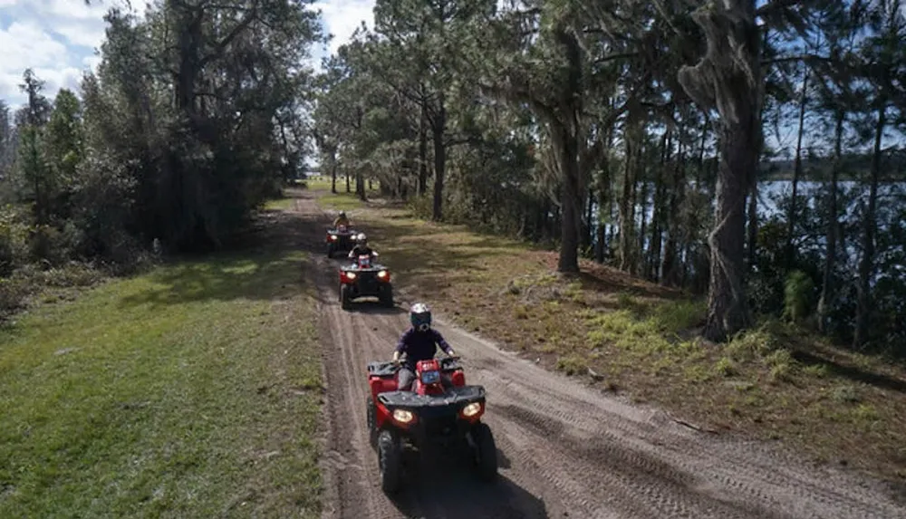 Four people are riding ATVs along a forested dirt path beside a lake