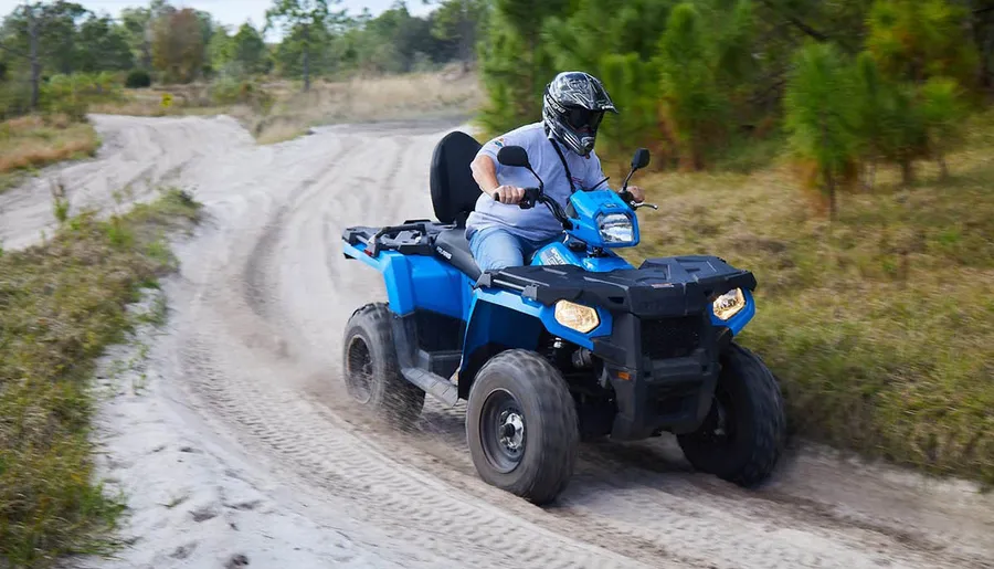 A person wearing a helmet is riding a blue ATV on a sandy trail surrounded by grass and trees.