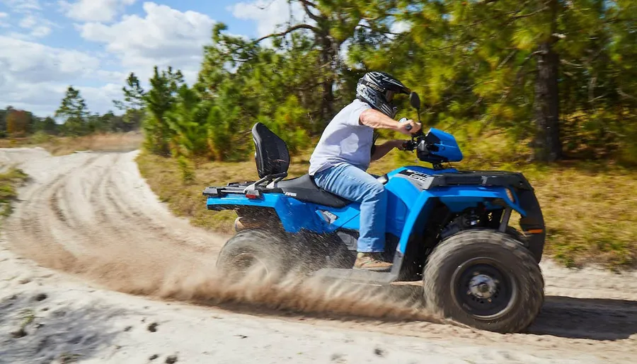 A person wearing a helmet is riding a blue ATV on a sandy trail surrounded by trees.