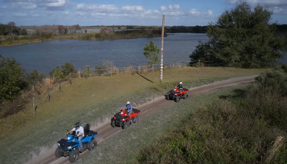 Three people ride ATVs on a dirt path next to a scenic lakeside area
