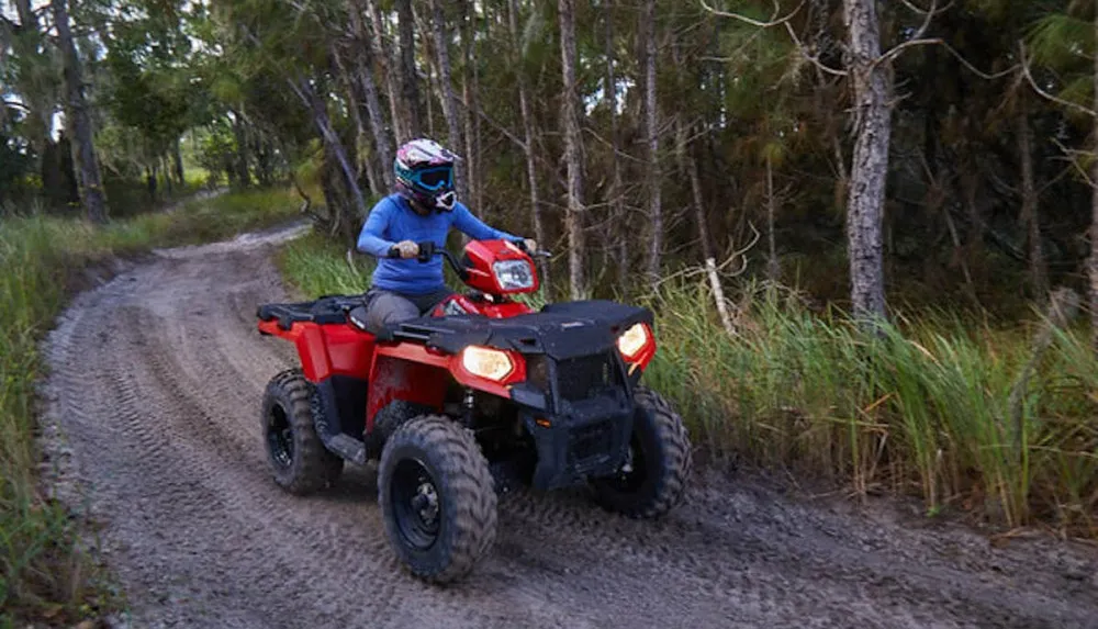 A person wearing a helmet is riding a red ATV on a dirt path through a forested area