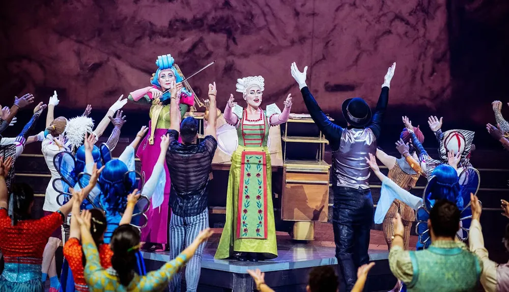A group of performers in colorful costumes and theatrical makeup are on stage enthusiastically raising their arms while a person in vibrant attire plays a violin