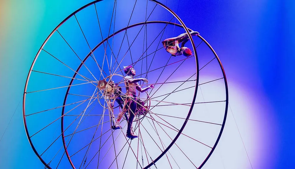 Performers are seen balancing on and within a large rotating wheel against a colorful vibrant background