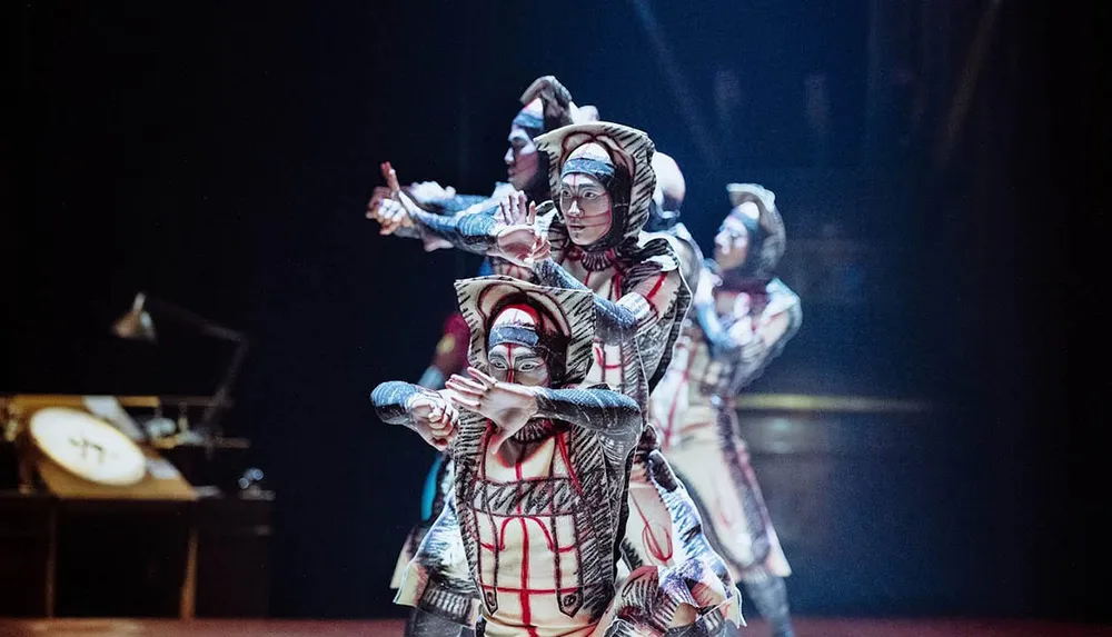 Four performers in elaborate costumes and face paint are striking synchronized poses on stage