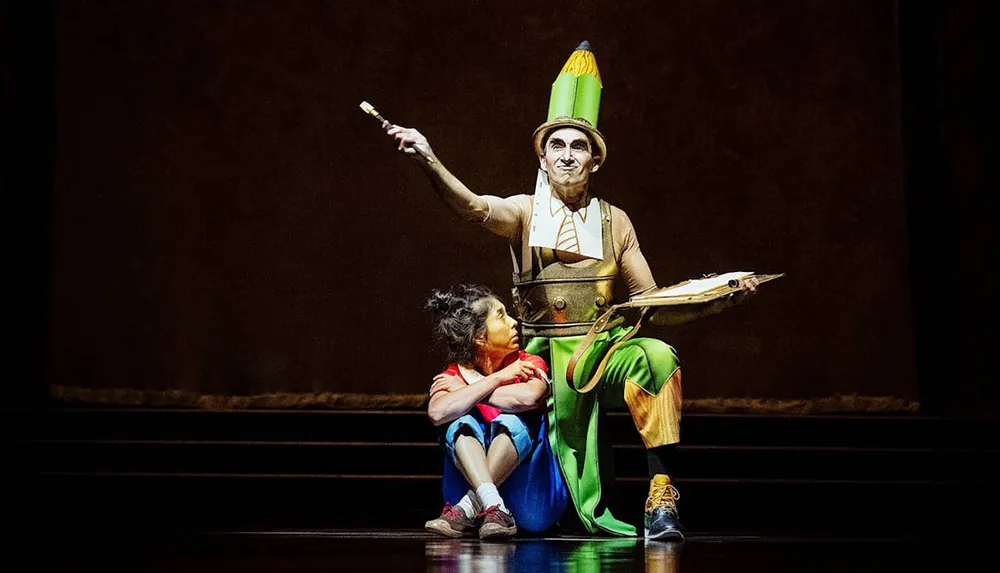 A performer dressed as a whimsical character with a pencil hat sits on stage next to a child-like figure holding an artists palette