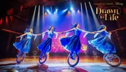 Four performers in blue costumes ride unicycles and hold hands on stage under bright lights, part of the 