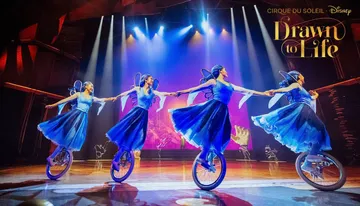 Four performers in blue costumes ride unicycles and hold hands on stage under bright lights, part of the 