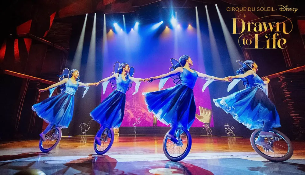 Four performers in blue costumes ride unicycles and hold hands on stage under bright lights part of the Drawn to Life Cirque du Soleil and Disney show