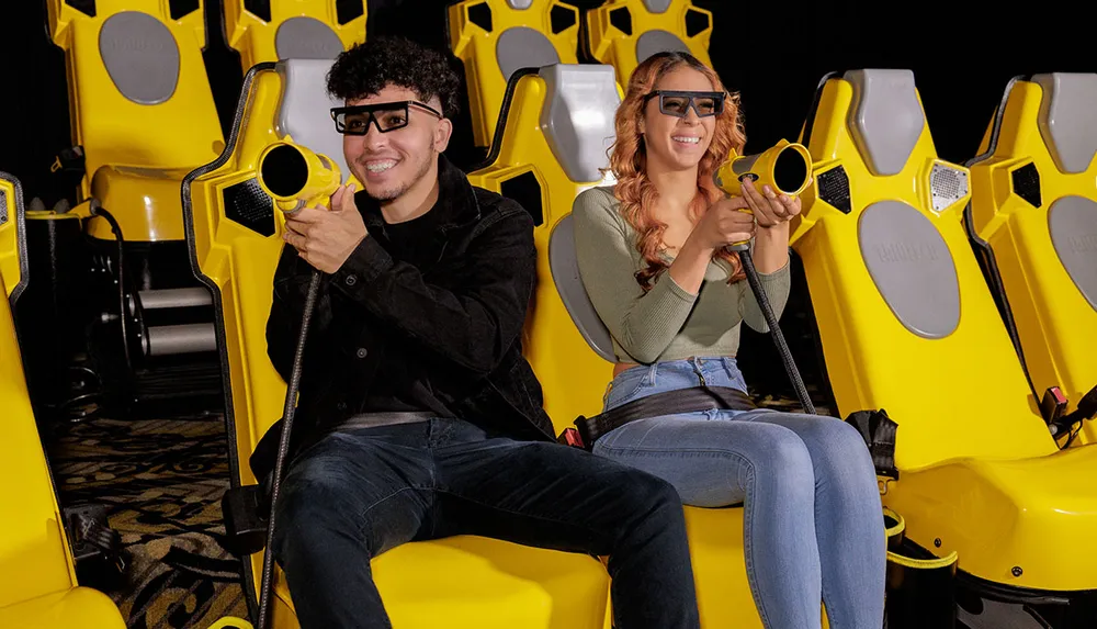 A couple is enjoying a 4D cinema experience sitting in yellow motion seats while wearing 3D glasses and holding interactive devices