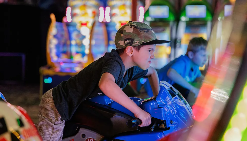 A young boy wearing a camouflage cap intensely rides a blue motorcycle arcade game with colorful lights in the background