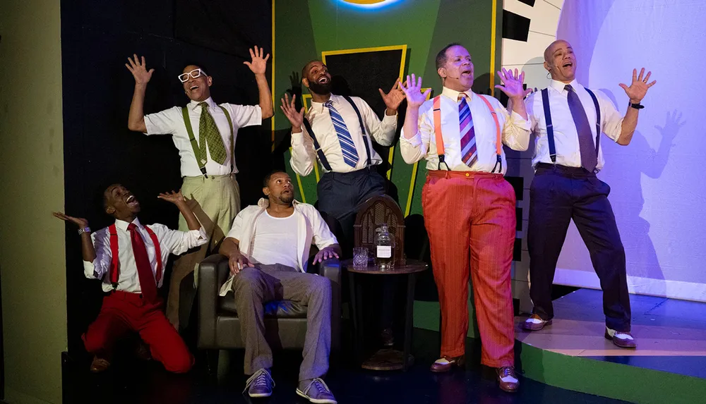 A group of six men dressed in colorful outfits with suspenders are energetically posing and gesturing on stage