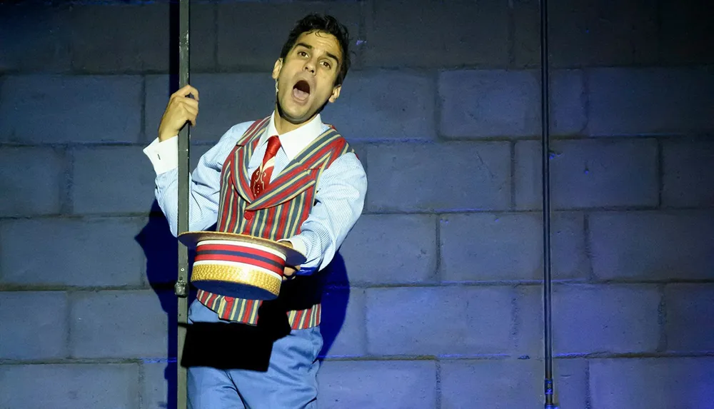 A performer in a striped vest and red tie sings passionately while holding a hat and a pole on stage