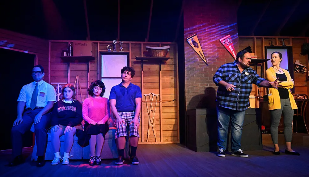 A group of people is performing a scene on stage with four seated and two standing in a setting resembling a room with wood-paneled walls and various props