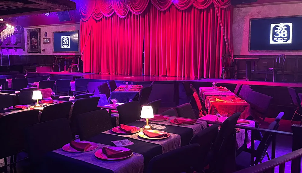 The image shows a dimly lit cabaret-style seating area with tables set up for dining facing a stage with red curtains