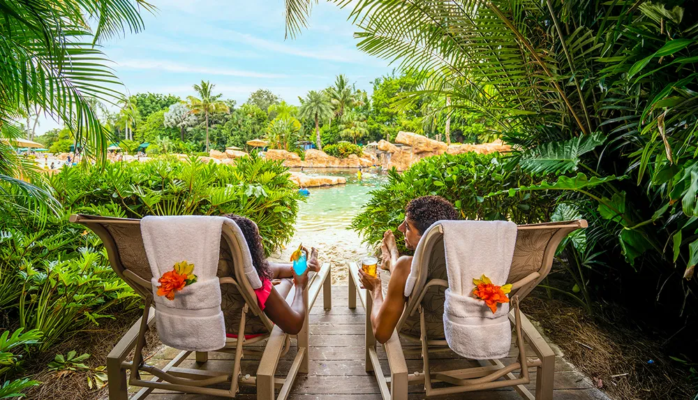 Two people relax on lounge chairs by a tropical pool surrounded by lush greenery enjoying colorful drinks