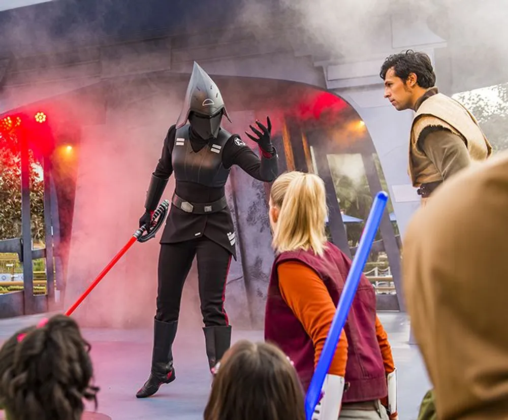 A performer in a dark costume with a red lightsaber is confronting a group of children with lightsabers in what appears to be a themed stage performance