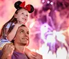 A joyful child sits on an adults shoulders enjoying a fireworks display while wearing Mickey Mouse ears