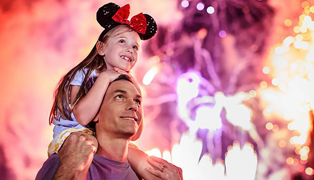 A joyful child sits on an adults shoulders enjoying a fireworks display while wearing Mickey Mouse ears