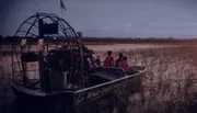 An airboat carrying passengers glides through a wetland area at twilight.