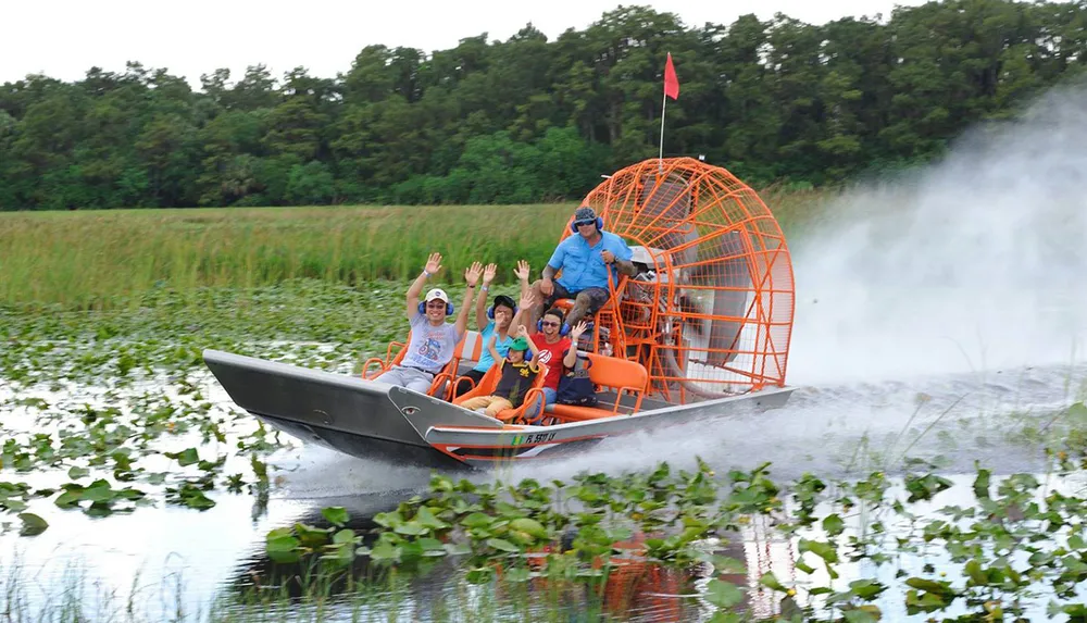 A group of people are joyfully waving while riding an airboat through a waterway with lush greenery on the sides