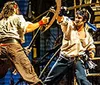 The image depicts a dynamic stage fight between two actors dressed as pirates superimposed with a smaller inset image of a plated meal featuring corn mashed potatoes and a piece of chicken