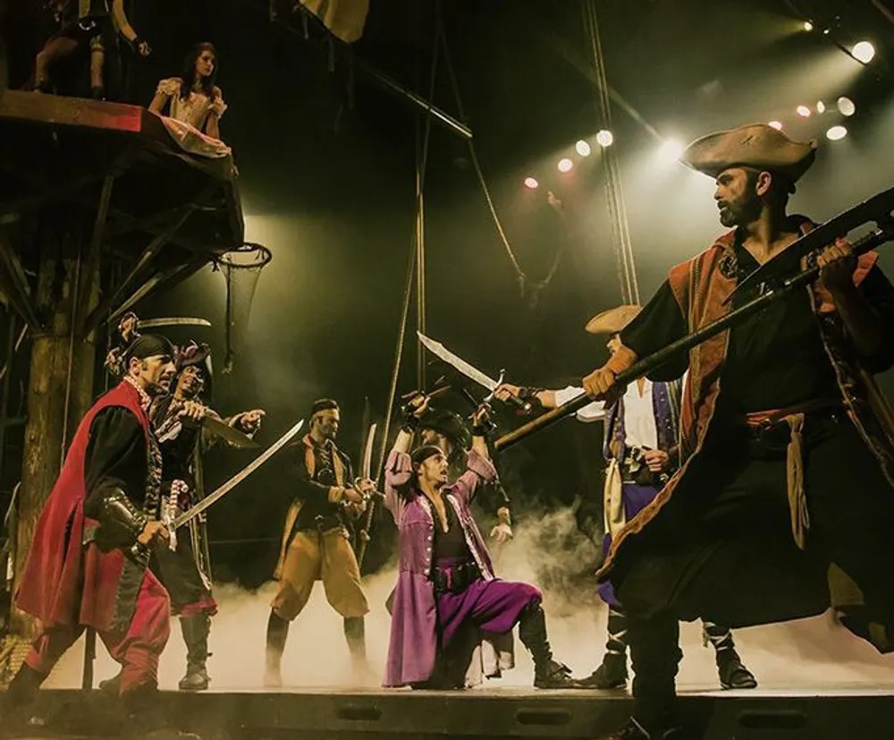 The image depicts a theatrical performance with actors in pirate costumes engaging in a dramatic sword fight on stage complete with atmospheric lighting and stage fog