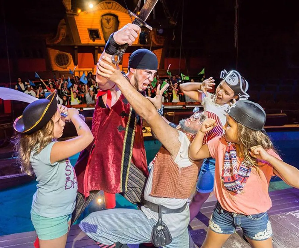 A group of children and performers dressed as pirates are engaged in a playful sword fight in the atmosphere of a ship-themed stage