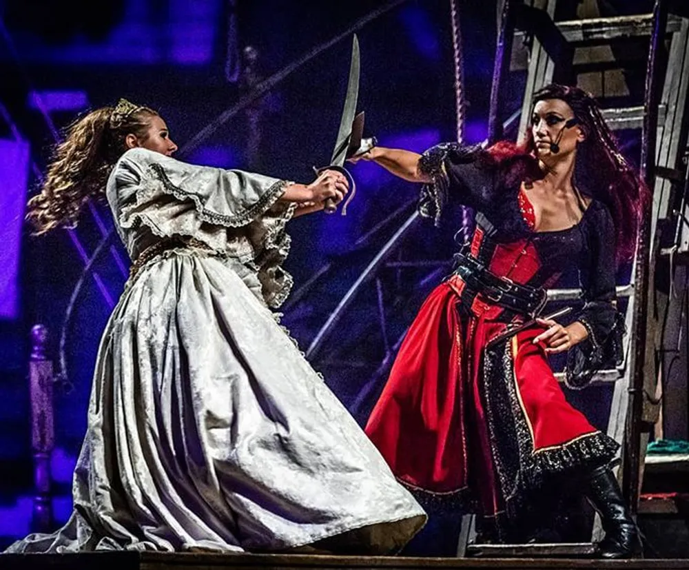 Two actors are engaged in a dramatic sword fight on a stage dressed in elaborate period costumes