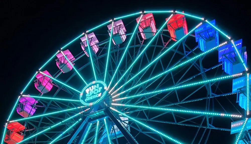 A brightly illuminated Ferris wheel at night features vibrant neon lights in a spectrum of blue to pink colors