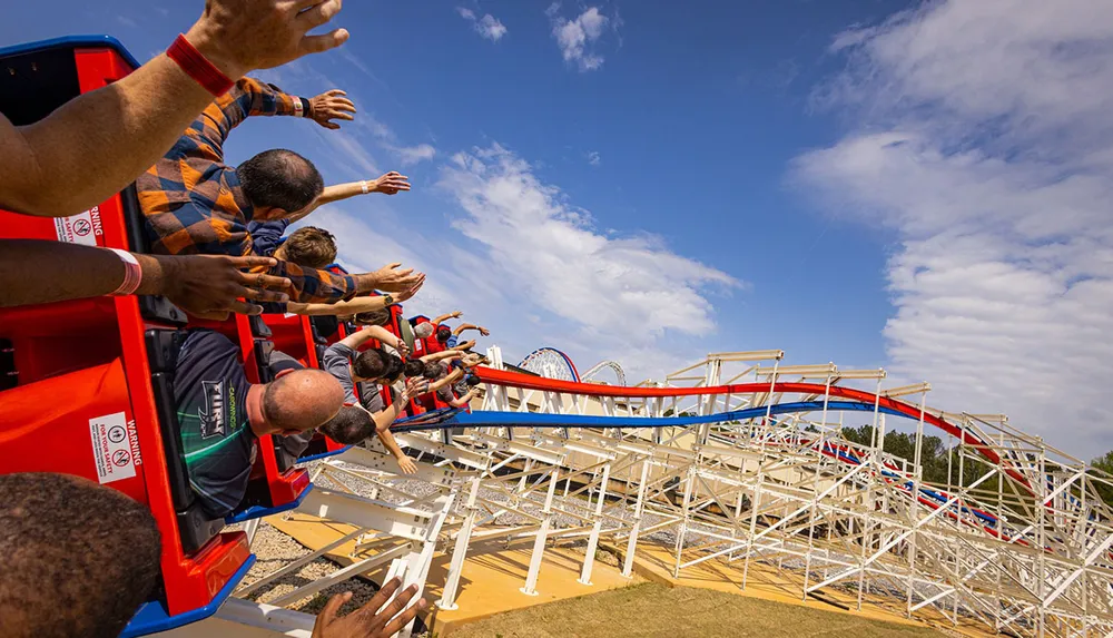 Thrill-seekers with their arms raised enjoy a lively descent on a colorful roller coaster under a blue sky dotted with clouds