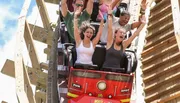 Thrilled passengers with raised arms are experiencing a steep drop on a wooden roller coaster under a clear sky.