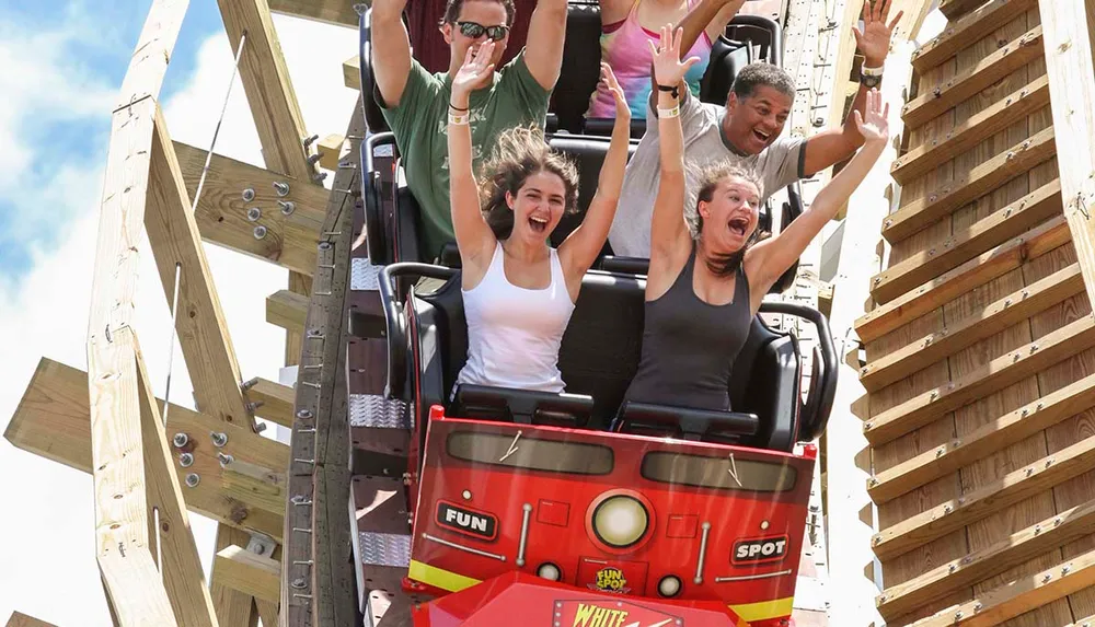 Thrilled passengers with raised arms are experiencing a steep drop on a wooden roller coaster under a clear sky