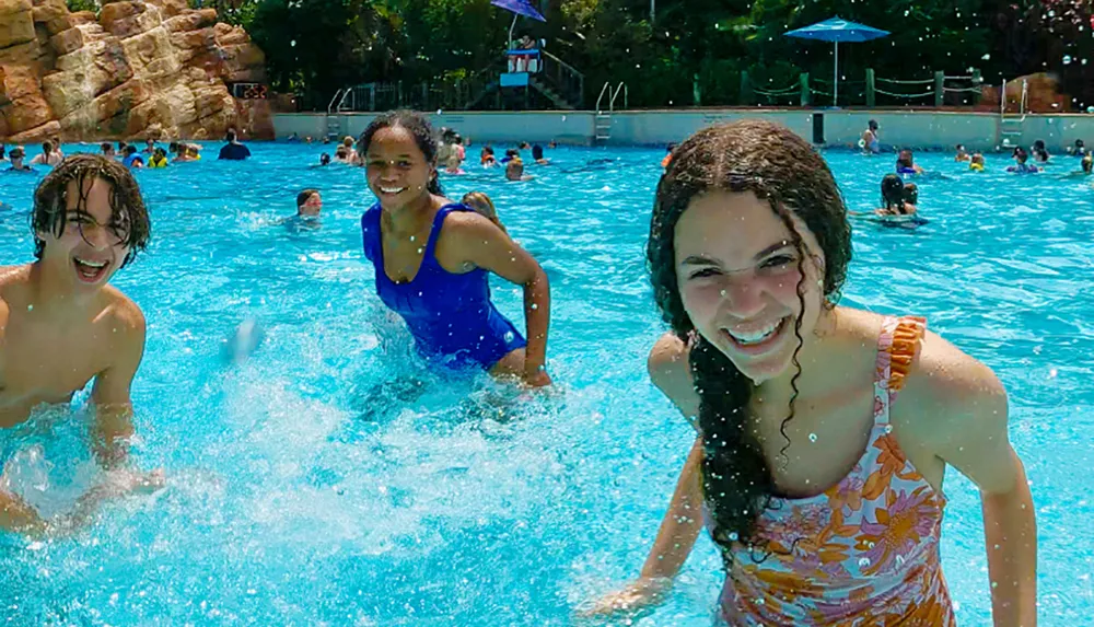 Three friends are joyfully splashing in a sunlit pool surrounded by other swimmers conveying a sense of summer fun