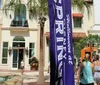 The image captures a sunny day at an outdoor shopping area with visitors walking past a banner palm trees and a small fountain