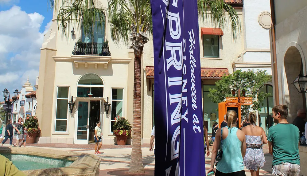 The image captures a sunny day at an outdoor shopping area with visitors walking past a banner palm trees and a small fountain