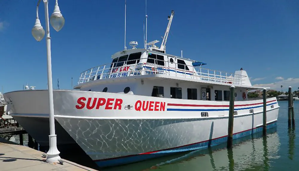 A large white and blue vessel named SUPER QUEEN is docked in a sunny marina with clear blue skies in the background