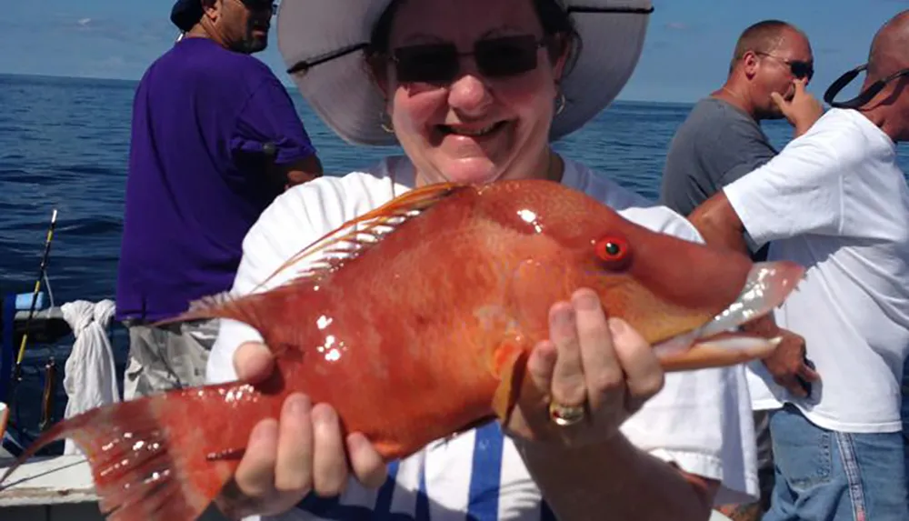 A smiling woman wearing a wide-brimmed hat proudly holds up a large orange fish aboard a boat with other people in the background