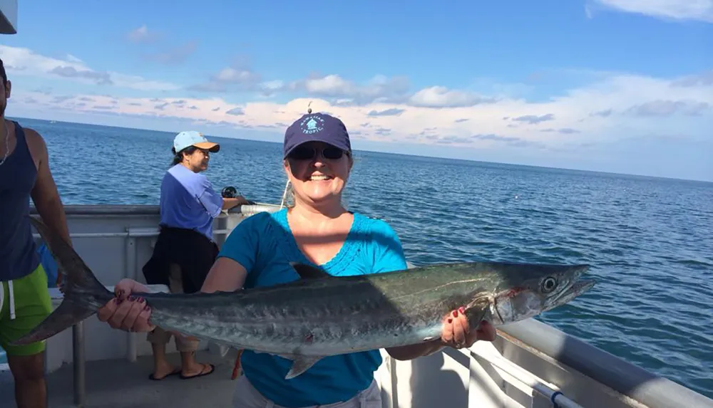 A woman is smiling for the camera while holding a large fish on a boat with other people around her set against a backdrop of calm sea and sky