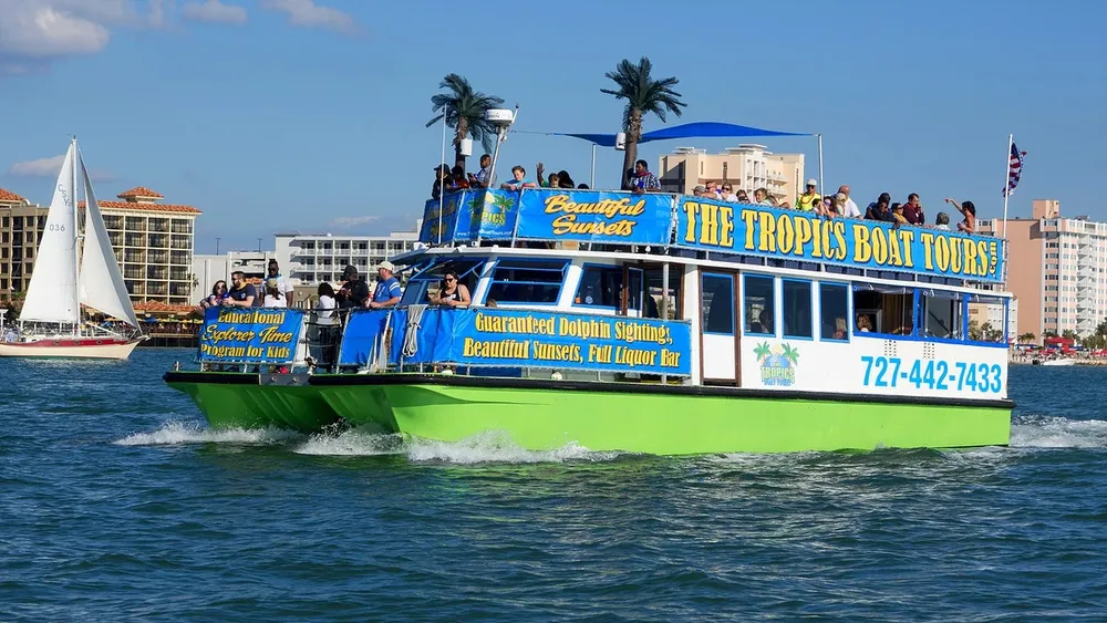 A colorful tourist boat promising dolphin sightings and beautiful sunsets is filled with passengers enjoying a trip on blue waters with a white sailboat and coastal buildings in the background