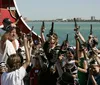 A red and black pirate-themed tour boat named Pirates Ransom is navigating a waterway with people on board with a bridge and palm trees in the background