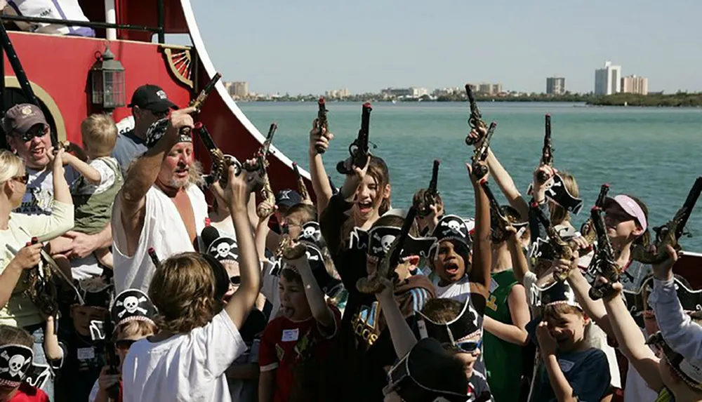 A group of excited children and adults some wearing pirate hats raise their toy guns in the air on a pirate-themed boat ride