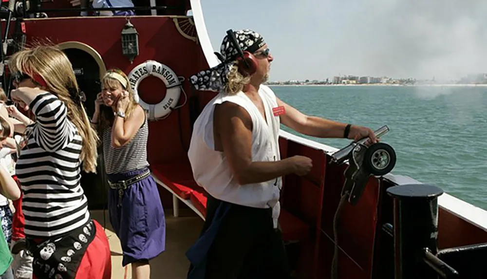 A person in pirate attire is steering a boat named Pirates Ransom with passengers looking on some covering their ears against a coastal backdrop
