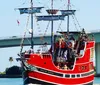 A red and black pirate-themed tour boat named Pirates Ransom is navigating a waterway with people on board with a bridge and palm trees in the background