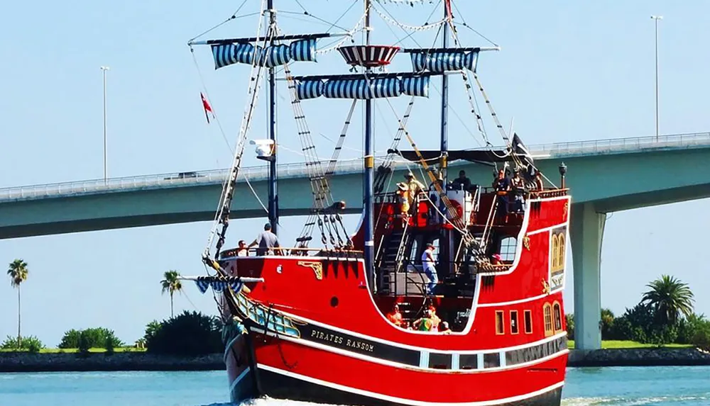 A red and black pirate-themed tour boat named Pirates Ransom is navigating a waterway with people on board with a bridge and palm trees in the background