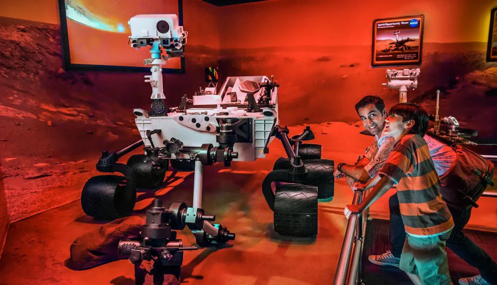 Two people are observing a Mars rover exhibit in a museum with a backdrop simulating the Martian surface