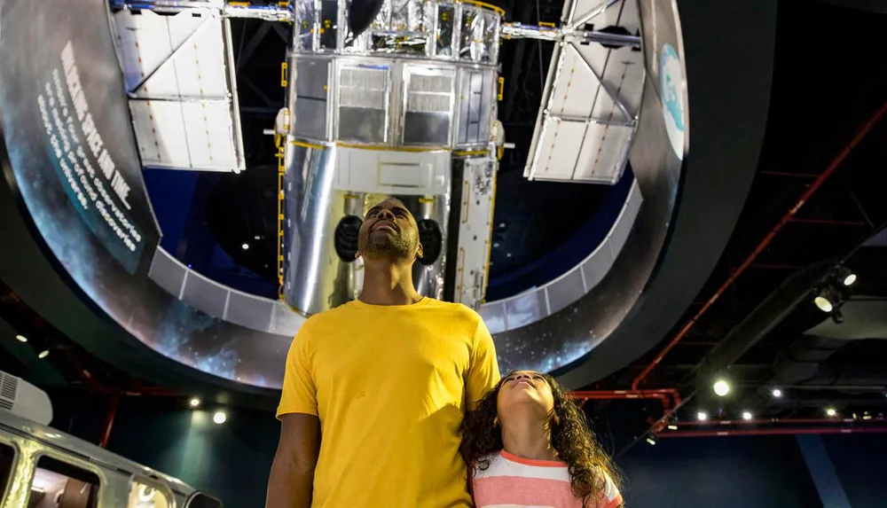 Two visitors are looking up at a large space telescope exhibit inside a museum