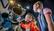 A boy in an astronaut costume holds a toy space shuttle with wide-eyed awe while a girl looks on, both in front of a backdrop of what appears to be a real spacecraft.