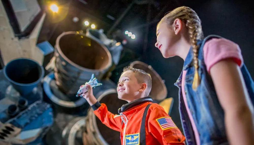A boy in an astronaut costume holds a toy space shuttle with wide-eyed awe while a girl looks on both in front of a backdrop of what appears to be a real spacecraft