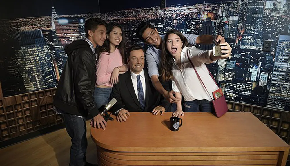 A group of enthusiastic individuals is taking a selfie with a figure at a talk show hosts desk set against the backdrop of a nighttime cityscape
