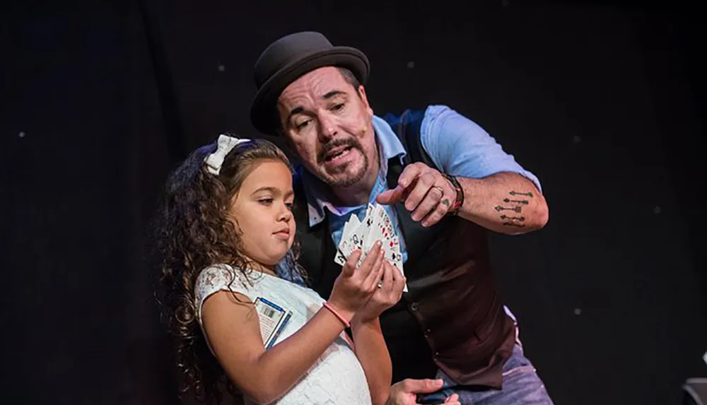 A magician wearing a hat is showing a card trick to a young girl on stage both looking at the cards shes holding